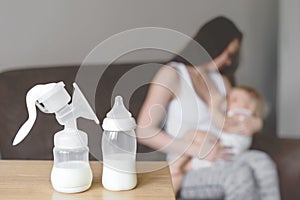 Bottle and breast pump with breast milk on the background of mother holding in her hands and breastfeeding baby. Maternity and bab