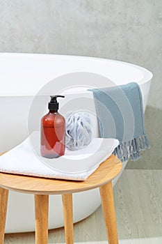 A bottle of brown shampoo and a bath sponge on a folded towel on a wooden table, next to a bathtub on a gray background. Space