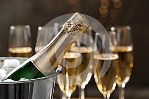 Bottle of champagne in bucket with ice and glasses on blurred background, closeup.