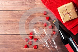 A bottle of champagne, two glass, a gift with a red ribbon, and red hearts flying are on a wooden background.