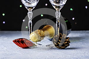 A bottle of champagne and two glass glasses with a pine cone and a Christmas tree toy next to the legs on a black background with