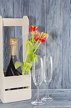 A bottle of champagne in a wooden box. next to 2 glasses of champagne and Tulip flowers. the holiday is March 8, mother`s Day.