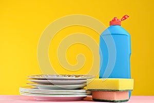 A bottle of dish washing, sponges, utensils on a wooden pastel color table against a yellow wall background, copy space.