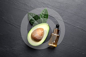 Bottle of essential oil, green leaves and fresh avocado on black table, flat lay