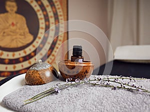 Bottle of essential oil in a round bowl of wood, next to a lavender branch, both arranged on a gray, rectangular terry towel, at