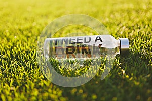 Bottle with fresh clean clear transparent water with text inscription I need drink on green grass on sunshine lawn