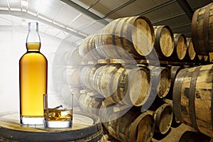 A bottle and glass of amber whisky set on top of an old barrel, in a barrel warehouse, back lit with light shinning through the