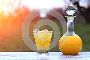 Bottle and glass with orange juice, ice cubes in glass at flower peach tree garden