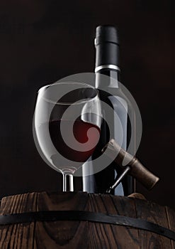 Bottle and glass of red wine with corks and vintage corkscrew on top of wooden barrel on black