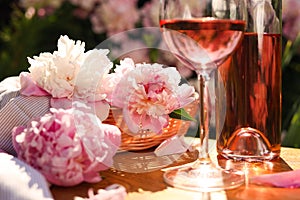 Bottle and glass of rose wine near beautiful peonies on wooden table in garden, closeup