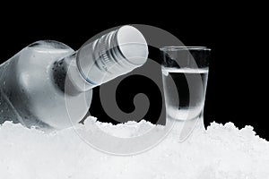 Bottle with glass of vodka lying on ice on black background
