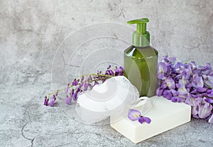 A bottle with liquid soap, a bar of white soap and natural flowers are on a gray background.