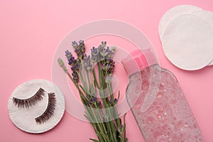 Bottle of makeup remover, lavender, cotton pads and false eyelashes on pink background, flat lay