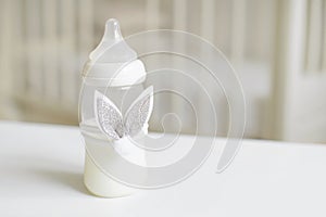 Bottle of milk for baby decorated with toy bunny`s ears over blurred background in children`s room. Free copy space.