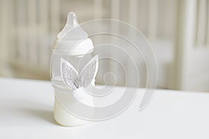 Bottle of milk for baby decorated with toy bunny`s ears over blurred background in children`s room. Free copy space. photo