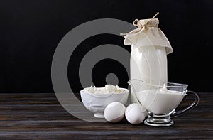 Bottle of milk, a plate of cottage cheese, a gravy boat with sour cream on a dark wooden background ,  raw eggs