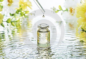 The bottle of Moisturizing cosmetic oil in the water waves on the summer flowers blur background and pipette with oil drop above