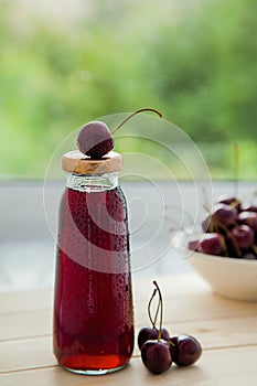 Bottle of natural cherry juice and white bowl with cherries on a wooden table near the window