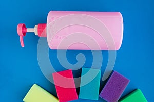 Bottle with pink dishwashing liquid and multicolor sponges on blue background.