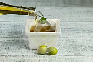 Bottle pouring virgin olive oil in white breakfast square bowl with raw green turkish olives on white rustic vintage wooden table.