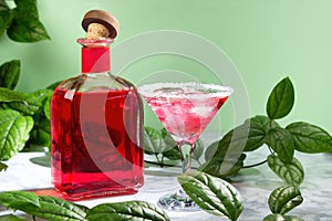 A bottle of red alcohol, next to a margarita glass is a cocktail with liquor and ice. On a marble and light green background