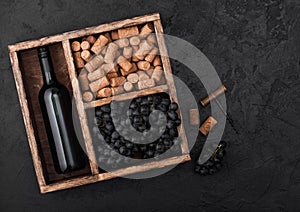 Bottle of red wine with dark grapes with corks and opener inside vintage wooden box on black stone background. Top view