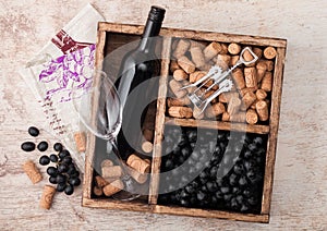 Bottle of red wine and empty glass with dark grapes with corks and opener inside vintage wooden box on wooden background with