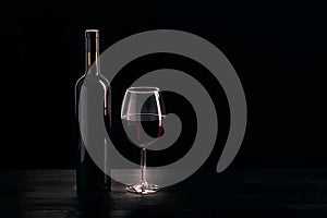 Bottle of red wine and a glass half filled with red wine, on a wooden black table, black background