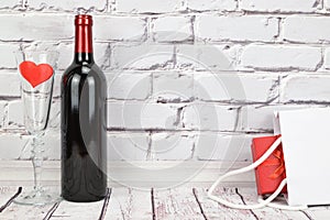 A bottle of red wine, a glass with a heart inside are on the wooden floor next to a white bag and a gift inside