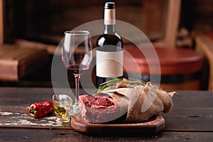 A bottle of red wine with a glass next to a piece of aged rib eye wrapped in parchment and burlap on a cutting board