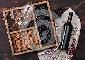Bottle of red wine on wood with empty glasses with dark grapes with corks and corkscrew inside vintage wooden box on dark wooden