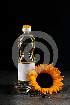 Bottle of sunflower cooking oil, seeds and beautiful flower on grey table against black background