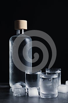 Bottle of vodka and shot glasses with ice on dark table against black background