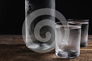 Bottle of vodka and shot glasses on wooden table against black background, closeup