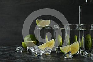 Bottle of vodka, shots, ice cubes and lime slices on black smokey table