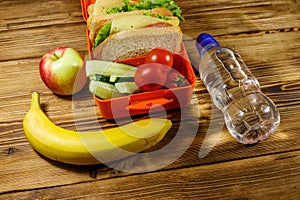 Bottle of water, apple, banana and lunch box with sandwiches and fresh vegetables on wooden table