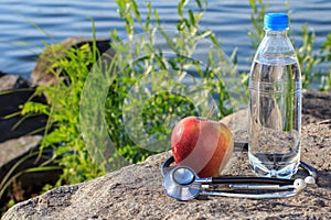 Bottle with water, apple and stethoscope on rock with river embankment background