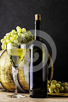 a bottle of wine a glass an oak barrel and a bunch of white grapes on a dark background