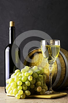a bottle of wine a glass an oak barrel and a bunch of white grapes on a dark background