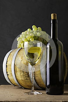 A bottle of wine a glass an oak barrel and a bunch of white grapes on a dark background