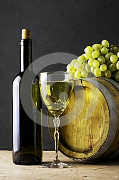 A bottle of wine a glass an oak barrel and a bunch of white grapes on a dark background