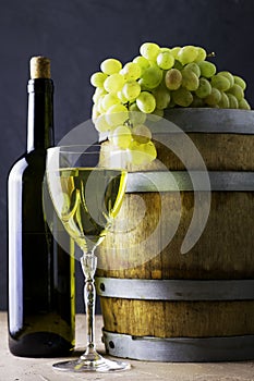 a bottle of wine a glass an oak barrel and a bunch of white grapes on a dark background