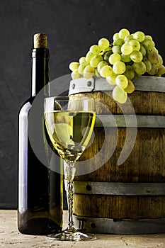 a bottle of wine a glass an oak barrel and a bunch of white grapes on a dark background