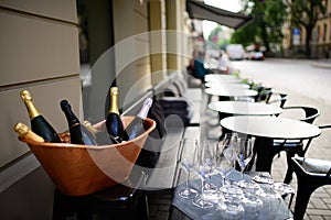 Bottles of champagne and sparkling wine in copper ice bucket and glasses nearby with row of empty tables and chairs on terrace of