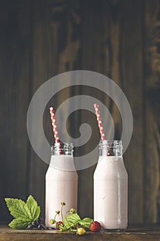 Bottles with delicious strawberry milkshake or smoothie with branch of wild strawberry on dark wooden table and background