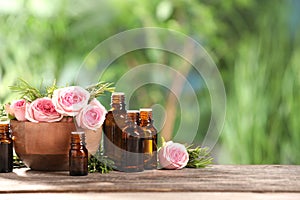 Bottles with essential oils, roses and rosemary on wooden table against blurred green background. Space for text