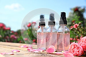 Bottles of facial toner with essential oil and fresh roses on wooden table against blurred background