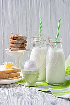 Bottles of milk with straws, boiled egg, toasts with butter and cookies, wooden background, healthy breakfast concept