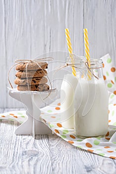 Bottles of milk with yellow striped straws and chocolate chip cookies on wooden background