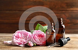 Bottles of rose essential oil, pipette and flowers on wooden table